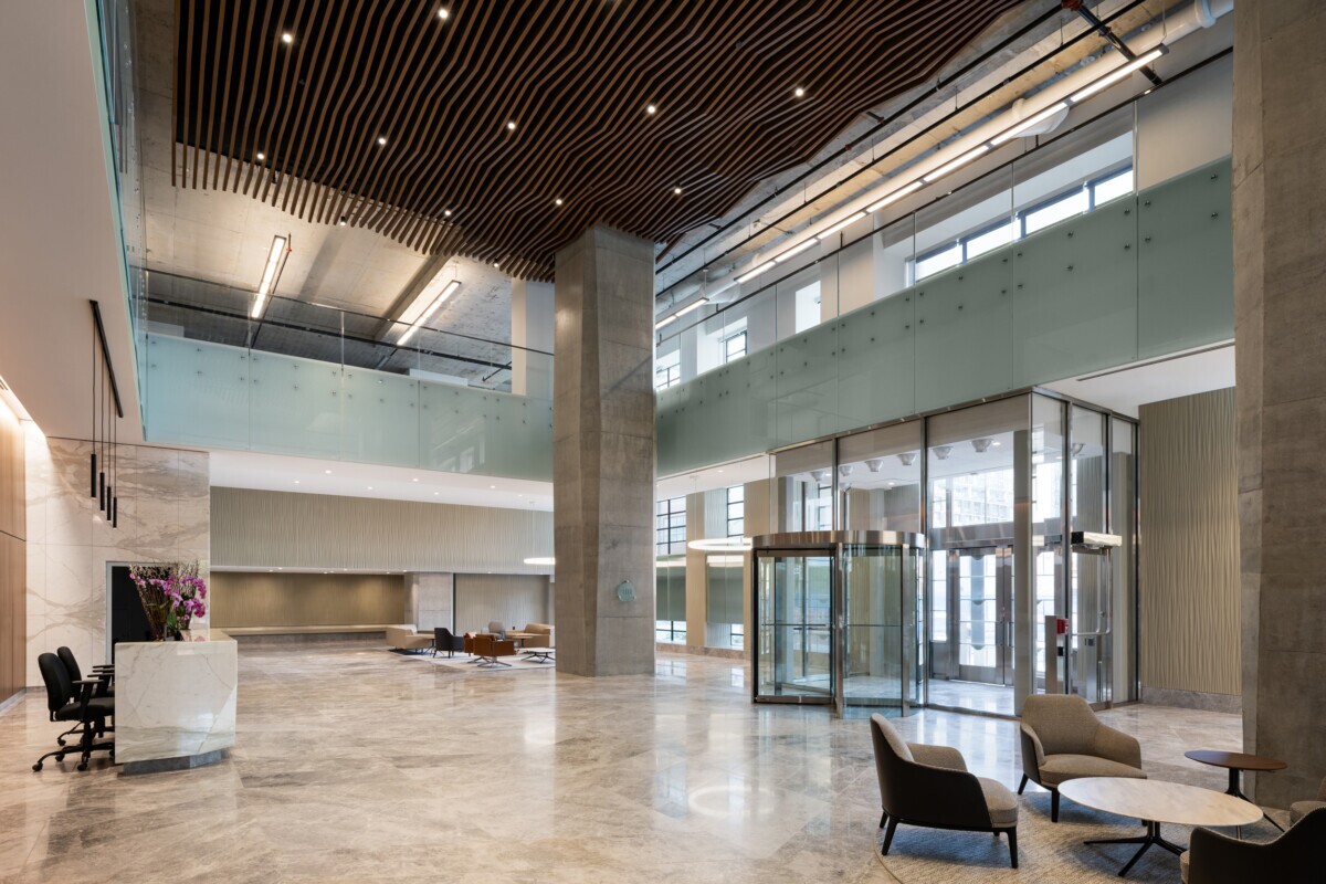 EQ Bank in Toronto, Canada atrium featuring a custom LINE:Form Wave Baffle ceiling feature in extruded metal that flows overhead. The ceiling’s undulating form brings a dynamic sense of movement and architectural expression to the open space. Finished in a warm wood veneer, the baffles offer a material counterpoint to the exposed cement walls, granite flooring, and structural pillars, softening the palette while maintaining a refined, modern aesthetic.