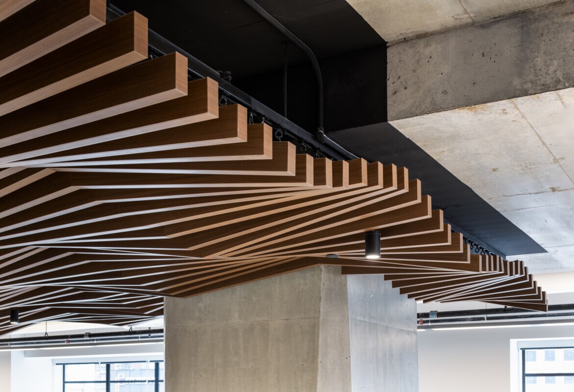 EQ Bank in Toronto, Canada close up of a custom LINE:Form Wave Baffle ceiling feature in extruded metal that flows overhead. The ceiling’s undulating form brings a dynamic sense of movement and architectural expression to the open space. Finished in a warm wood veneer, the baffles offer a material counterpoint to the exposed cement walls, granite flooring, and structural pillars, softening the palette while maintaining a refined, modern aesthetic.