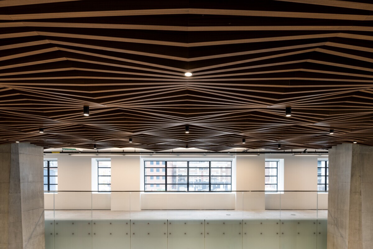 EQ Bank in Toronto, Canada atrium featuring a custom LINE:Form Wave Baffle ceiling feature in extruded metal that flows overhead. The ceiling’s undulating form brings a dynamic sense of movement and architectural expression to the open space. Finished in a warm wood veneer, the baffles offer a material counterpoint to the exposed cement walls, granite flooring, and structural pillars, softening the palette while maintaining a refined, modern aesthetic.