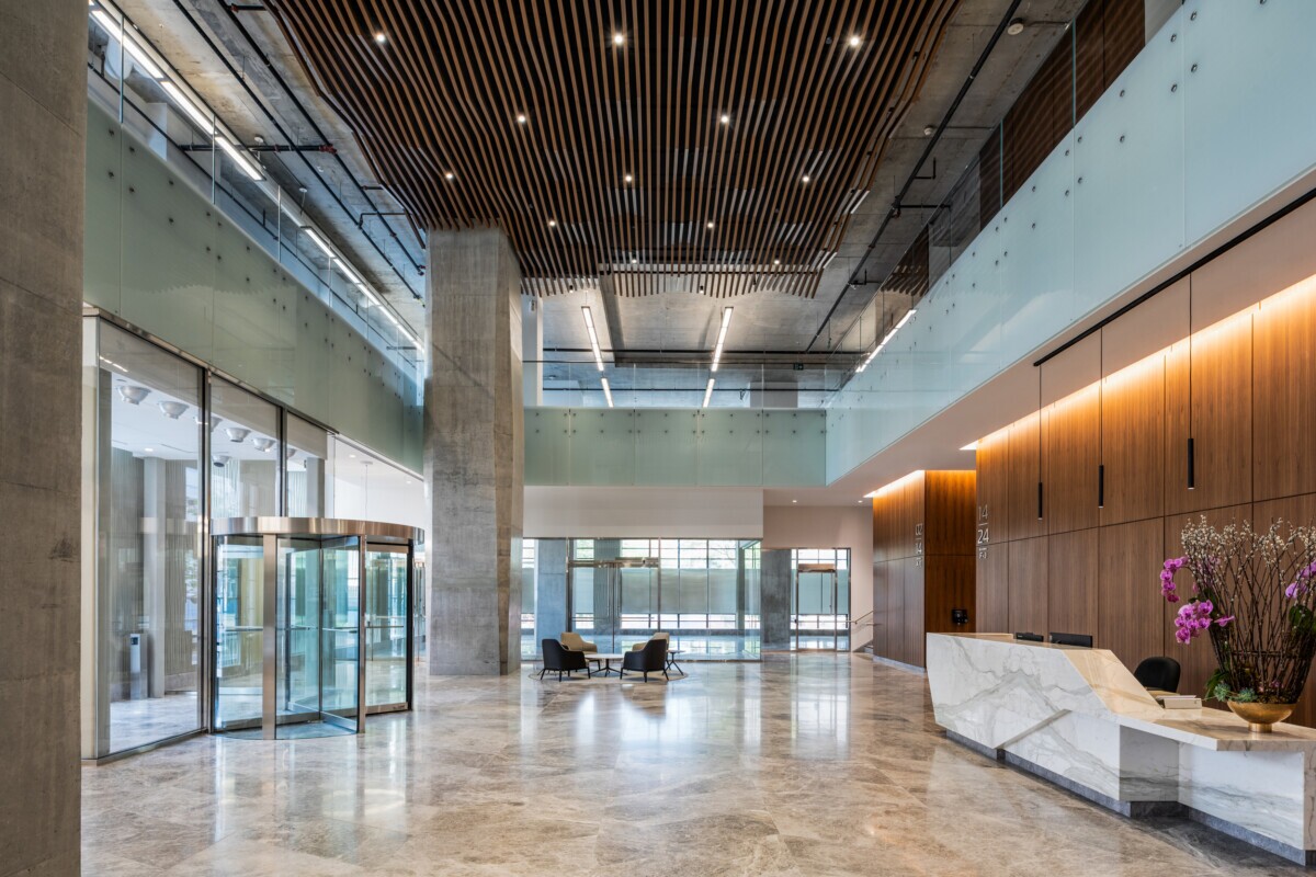 EQ Bank in Toronto, Canada atrium featuring a custom LINE:Form Wave Baffle ceiling feature in extruded metal that flows overhead. The ceiling’s undulating form brings a dynamic sense of movement and architectural expression to the open space. Finished in a warm wood veneer, the baffles offer a material counterpoint to the exposed cement walls, granite flooring, and structural pillars, softening the palette while maintaining a refined, modern aesthetic.