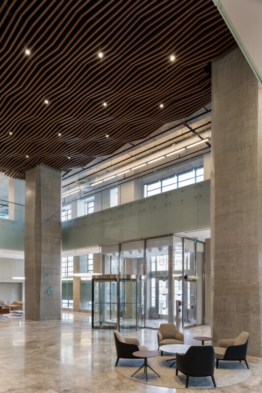 EQ Bank in Toronto, Canada atrium featuring a custom LINE:Form Wave Baffle ceiling feature in extruded metal that flows overhead. The ceiling’s undulating form brings a dynamic sense of movement and architectural expression to the open space. Finished in a warm wood veneer, the baffles offer a material counterpoint to the exposed cement walls, granite flooring, and structural pillars, softening the palette while maintaining a refined, modern aesthetic.