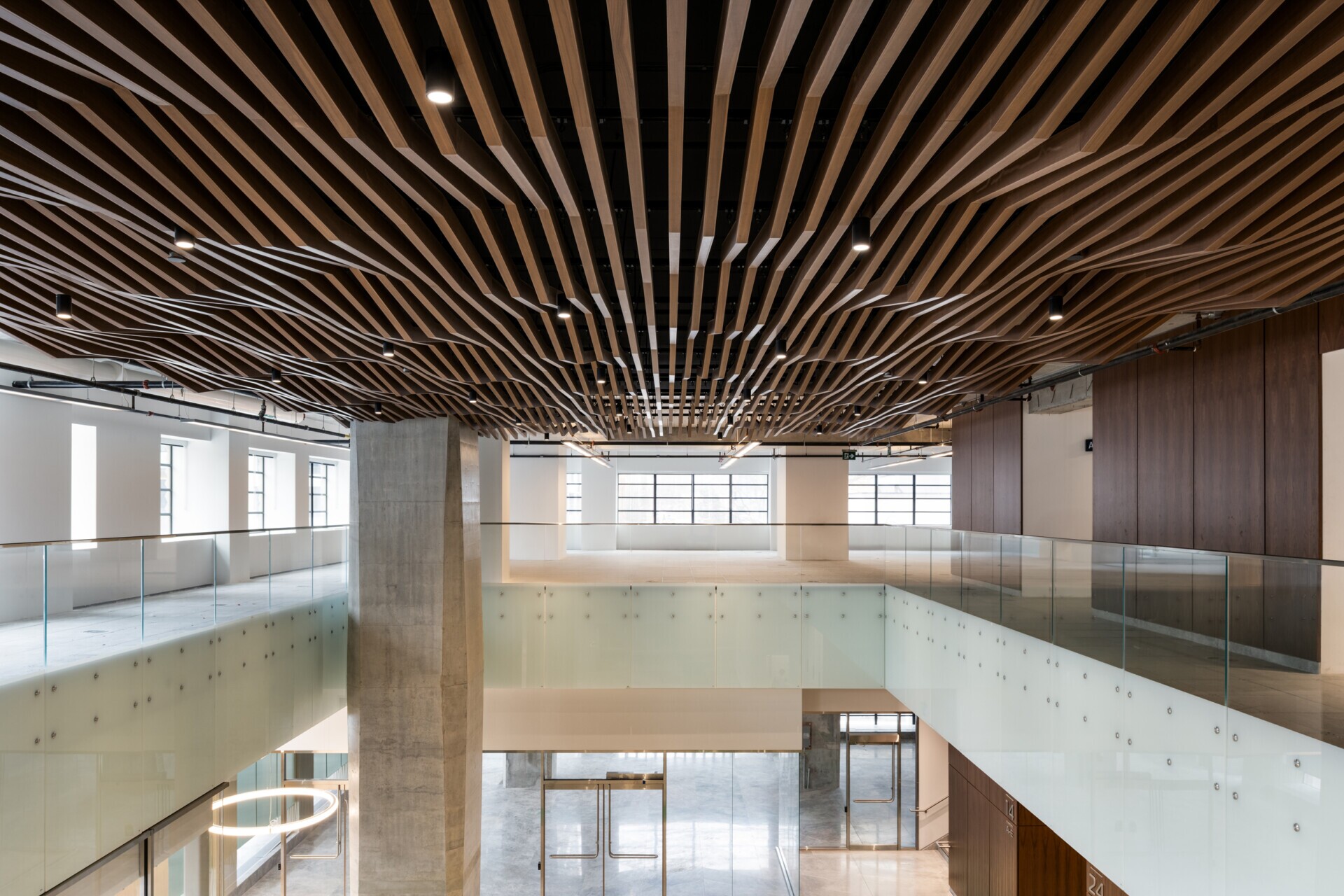 EQ Bank in Toronto, Canada atrium feautring a custom LINE:Form Wave Baffle ceiling feature in extruded metal that flows overhead.The ceiling’s undulating form brings a dynamic sense of movement and architectural expression to the open space. Finished in a warm wood veneer, the baffles offer a material counterpoint to the exposed cement walls, granite flooring, and structural pillars, softening the palette while maintaining a refined, modern aesthetic.